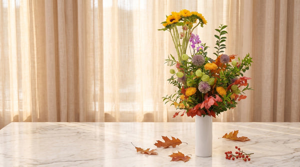 Tall autumn arrangement with sunflowers, allium and fiery fall foliage in a white vase, with fallen oak leaves and red berries on a marble table