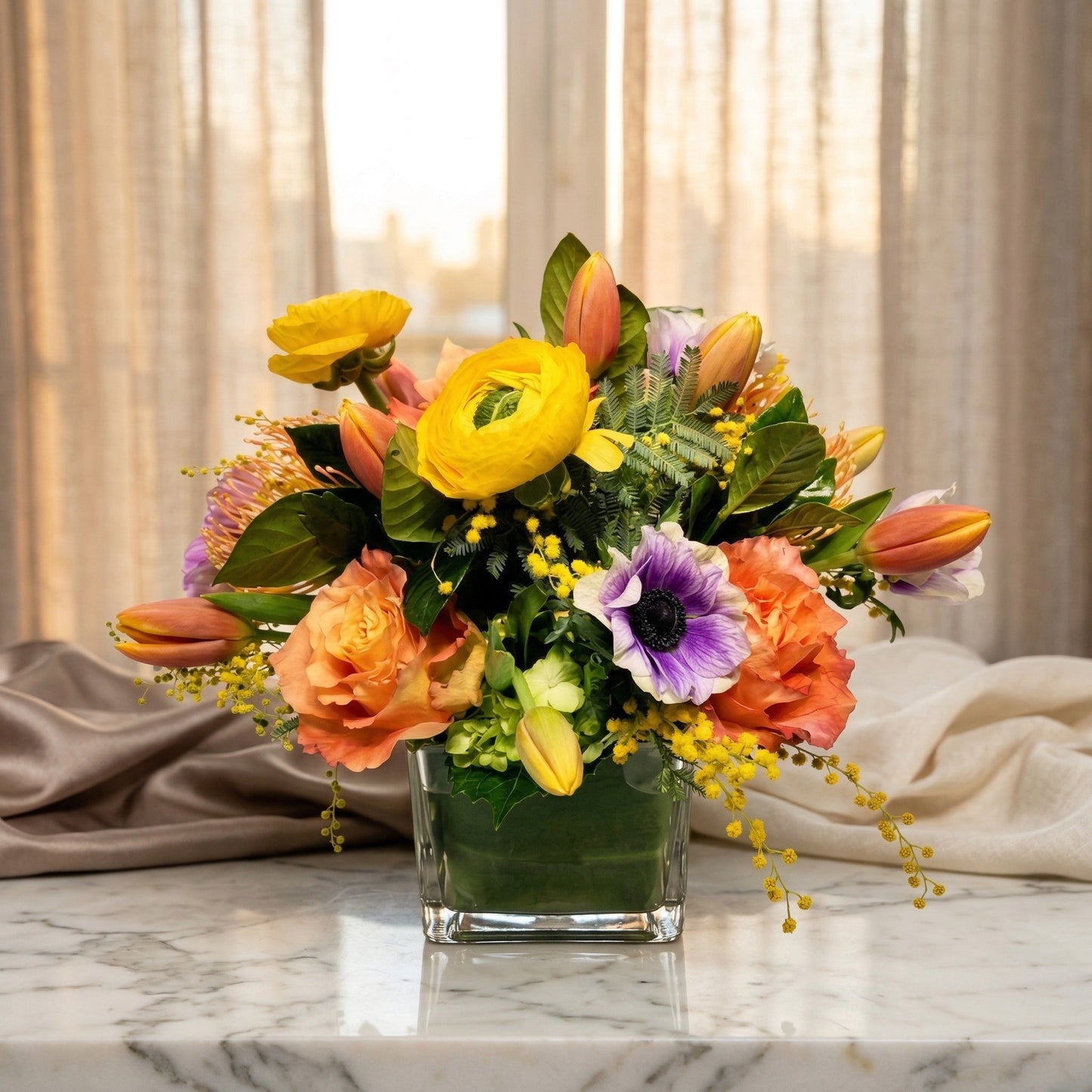 A vibrant mixed bouquet with yellow ranunculus, orange roses, and purple anemone in a glass vase - TJ Flowers NYC