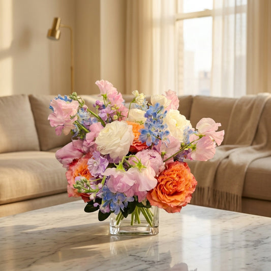 Mixed pastel bouquet in a glass vase featuring orange roses, white ranunculus, and delicate pink and blue blooms - TJ Flowers NYC
