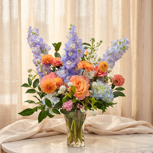 Tall arrangement of lavender delphinium with orange roses, orange dahlias and pale blue hydrangea in a clear glass vase - TJ Flowers NYC