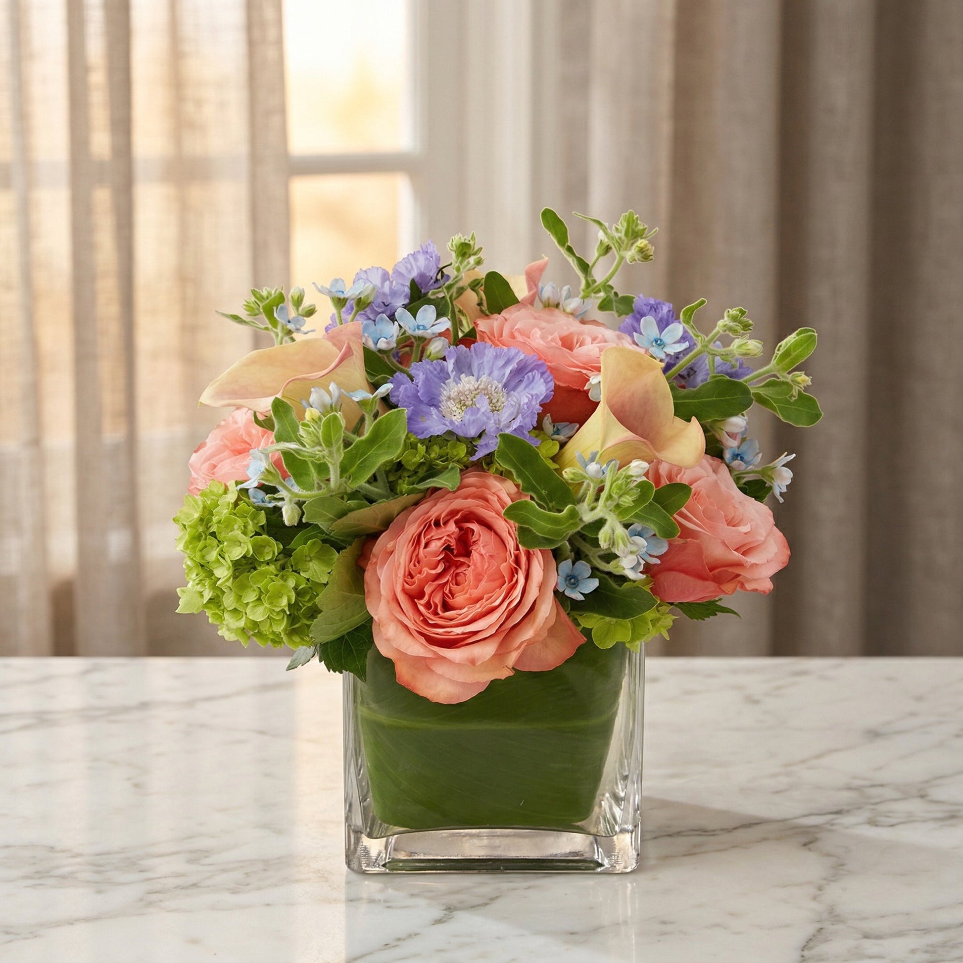 Pastel peach roses, blue scabiosa, and green hydrangea in a leaf-lined square glass vase - TJ Flowers NYC