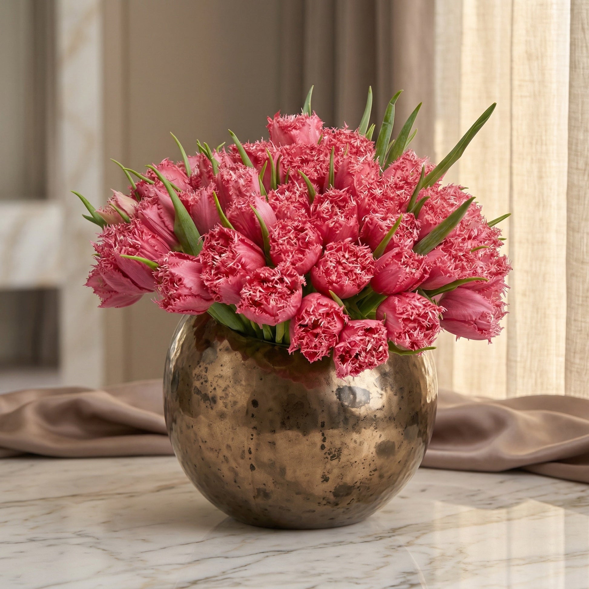 A large bouquet of vibrant pink fringed tulips in a round, textured bronze vase on a marble table. - TJ Flowers NYC