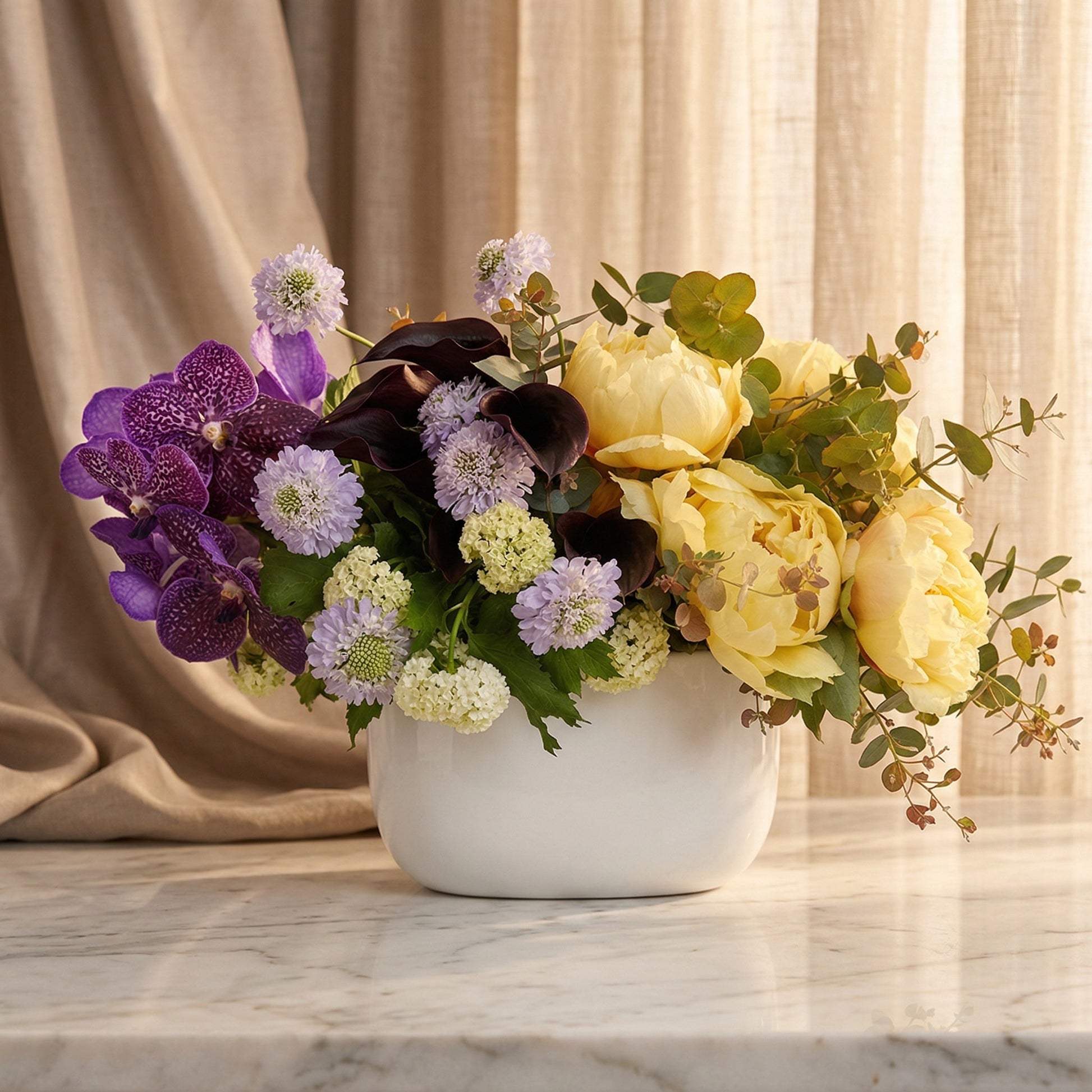 Pastel yellow peonies, purple orchids, and scabiosa in a white ceramic vase - TJ Flowers NYC