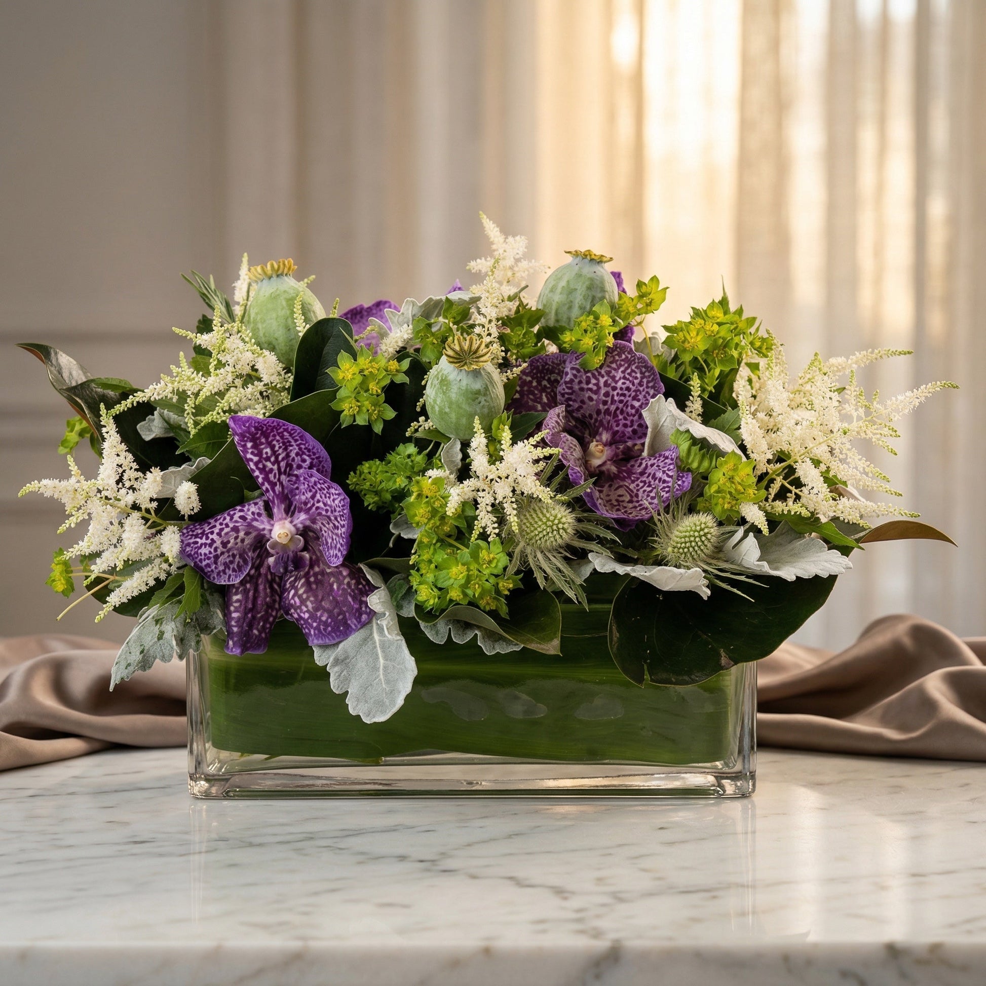 Purple Vanda orchids, white astilbe, and green poppy pods in a clear glass rectangular vase - TJ Flowers NYC
