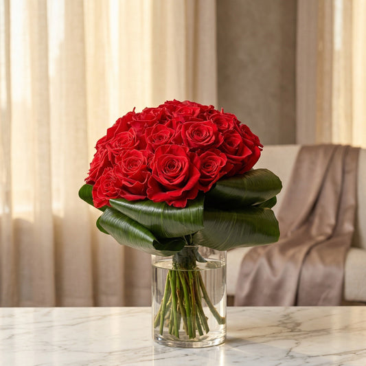 A large bouquet of deep red roses with large green leaves in a clear glass vase. - TJ Flowers NYC