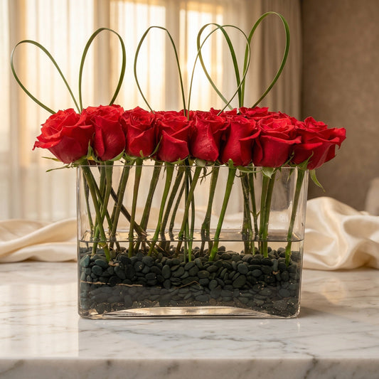Modern arrangement of red roses in a clear rectangular vase with black pebbles and grass loops - TJ Flowers NYC