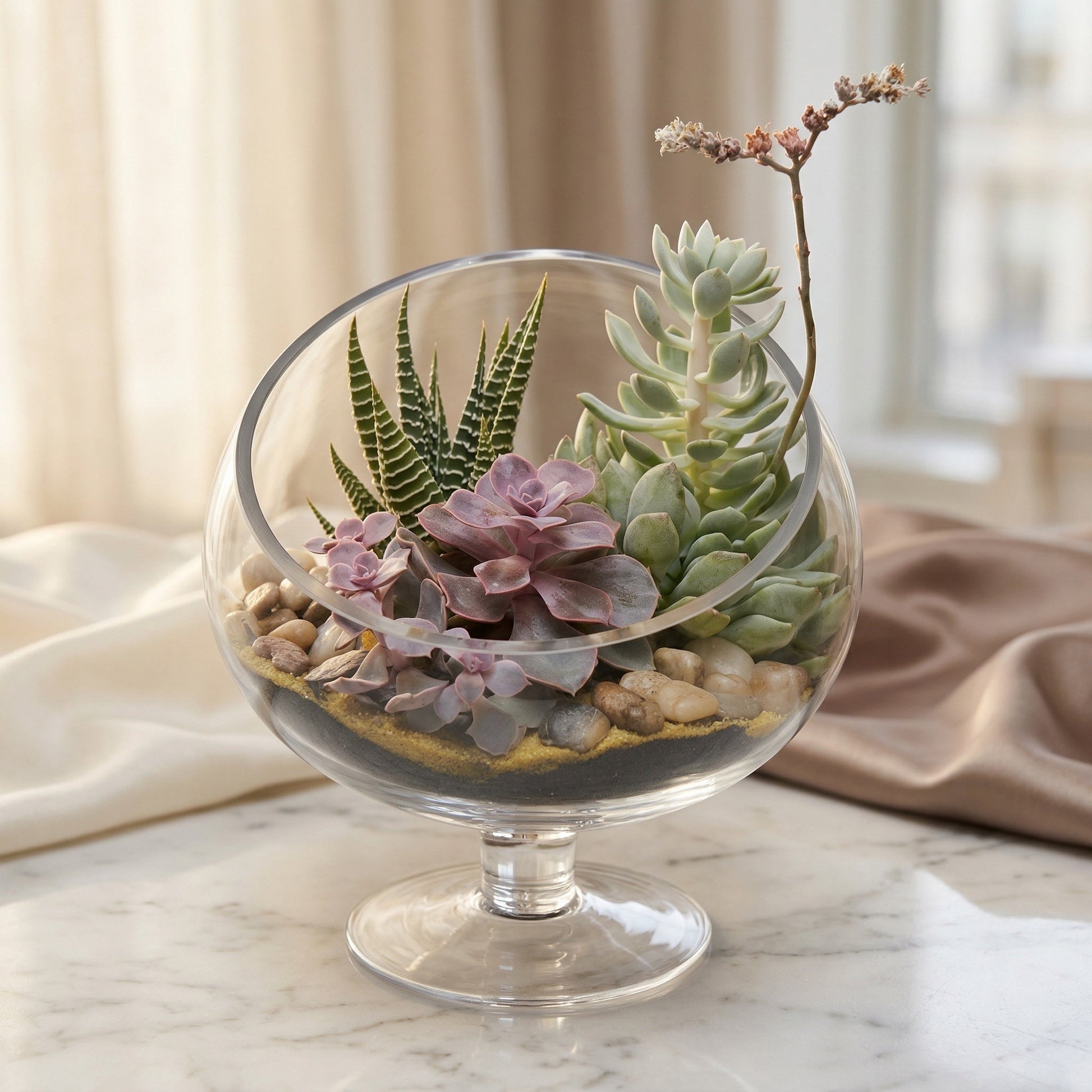 A glass terrarium with purple and green succulents, sand, and pebbles on a marble table. - TJ Flowers NYC