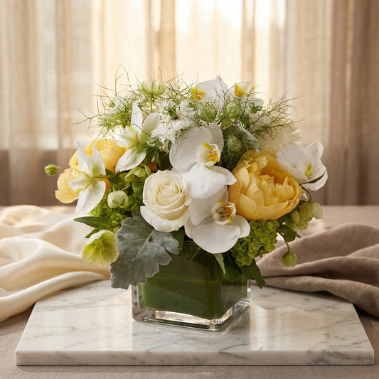 White orchids, yellow peonies, and roses in a square glass vase with green foliage - TJ Flowers NYC