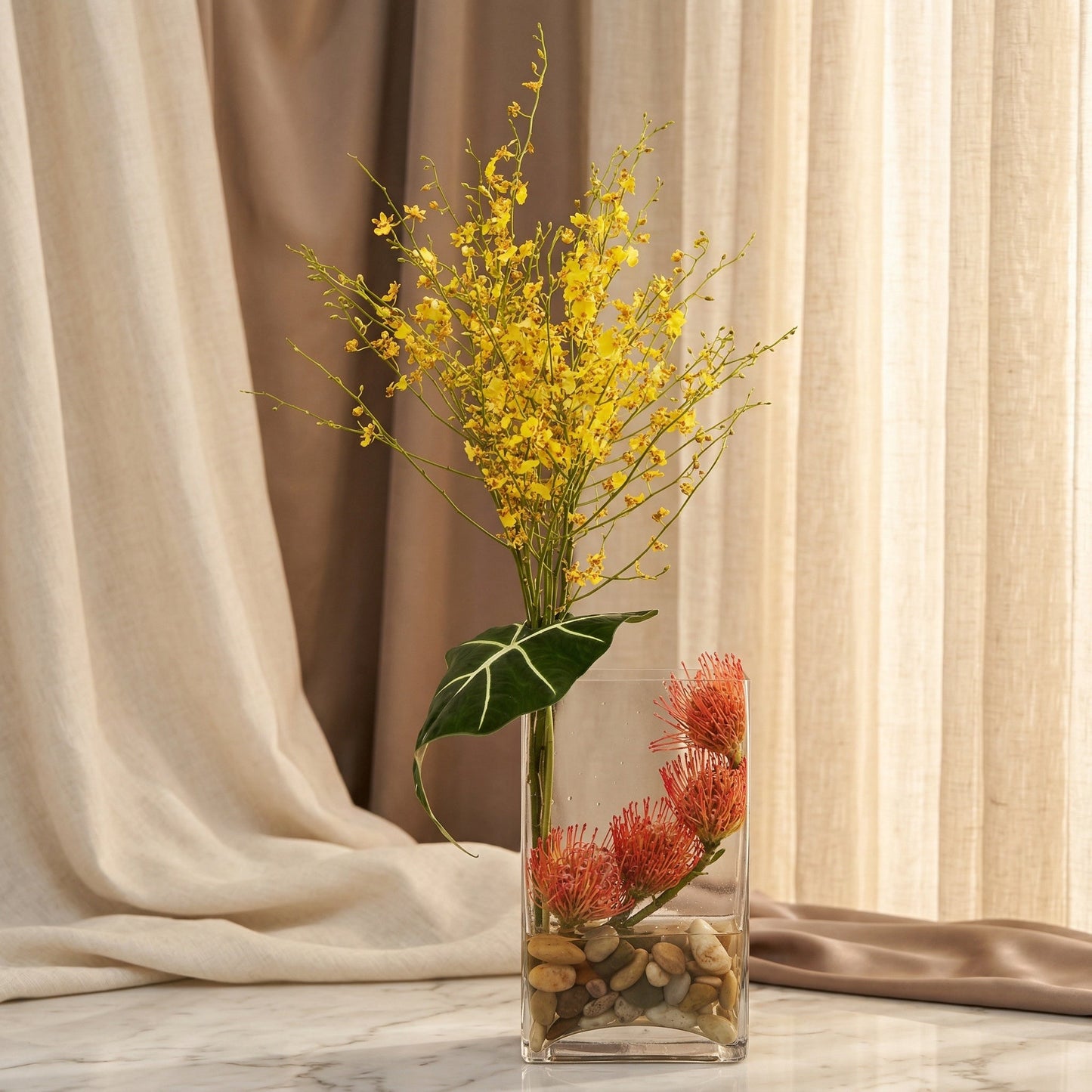 Yellow oncidium orchids and orange pincushion protea in a clear glass vase with river rocks - TJ Flowers NYC