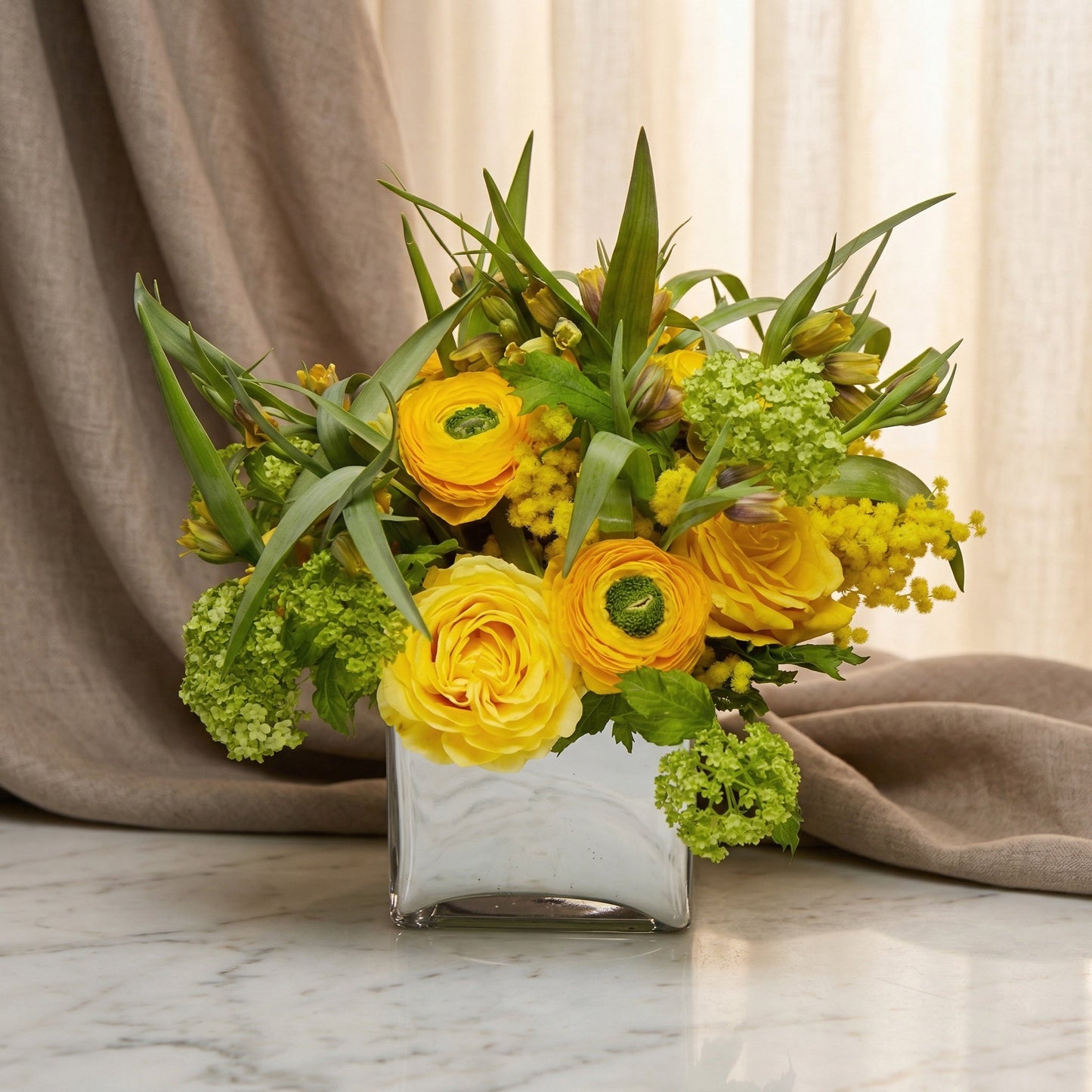 Yellow roses, ranunculus, and green viburnum in a square silver vase. - TJ Flowers NYC