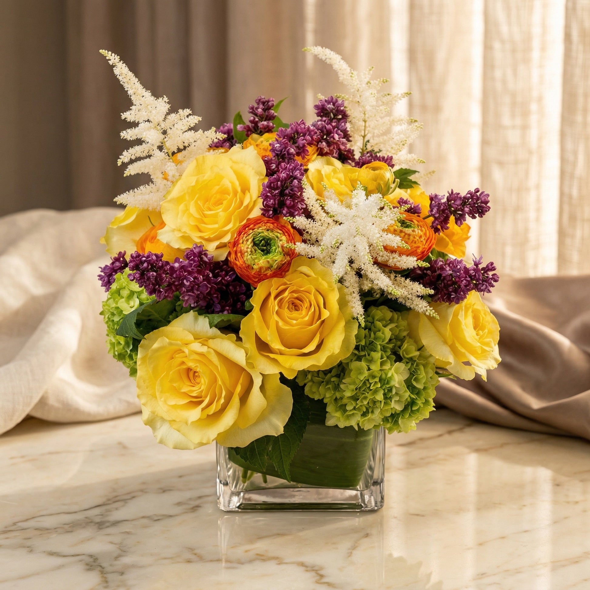 Yellow roses, purple lilac, and green hydrangeas in a square glass vase. - TJ Flowers NYC